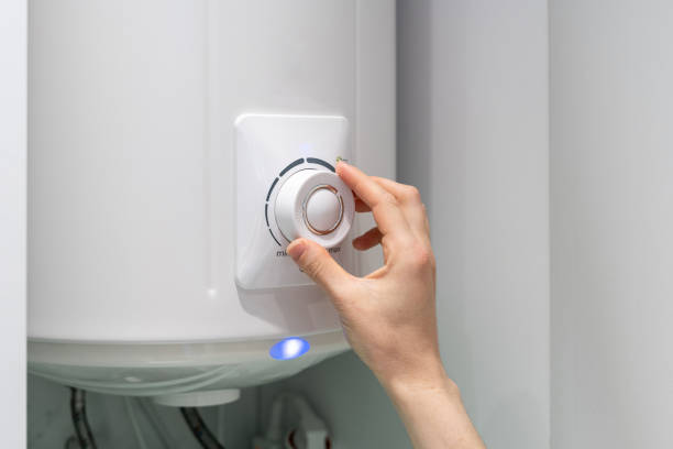 A right hand can be seen adjusting the temperature dial on a water heater.