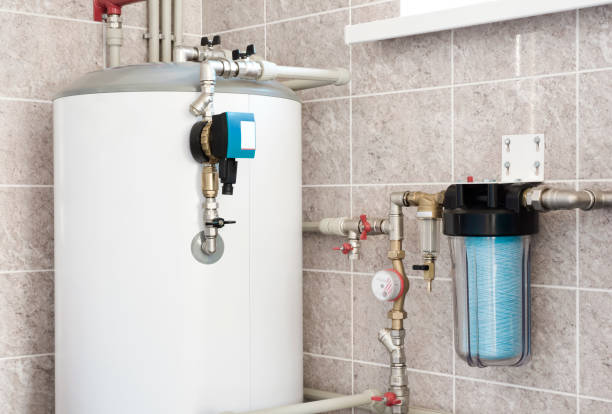 A residential water heating boiler with pump, ball valves and filters sits in the corner of a room with beige tile walls.