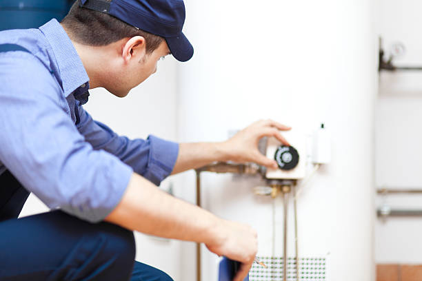 A plumbing technician wearing a blue hat and button up shirt adjusts the dial on a hot water heater while crouching down.